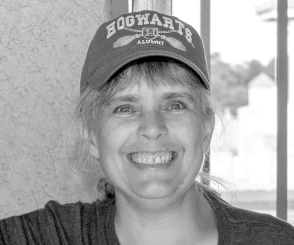 A smiling woman in a Hogwarts Alumni cap by a window